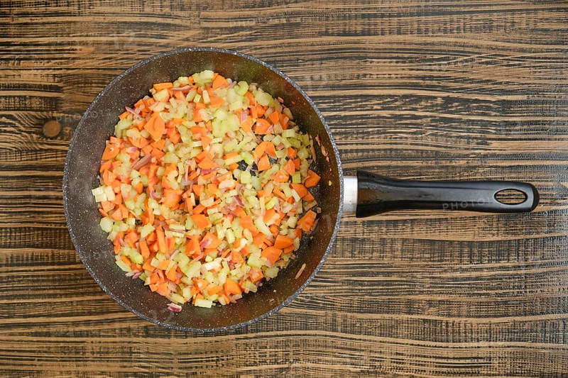 Colorful chopped carrots, onions, and celery are cooking in a black skillet on a wooden table. The vibrant mix of vegetables is ready to be used in a delicious recipe.