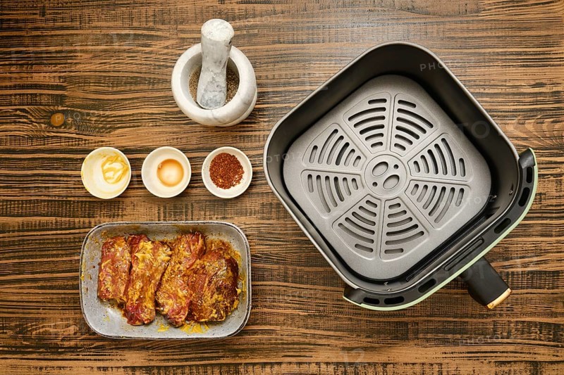 Marinated beef meat next to bowls of mustard, soy sauce, and spice blend on a wooden table. An air fryer sits ready for cooking, inviting delicious meal preparations