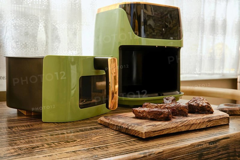 A stylish kitchen corner showcases an air fryer beside a cooking unit. Juicy pieces of grilled meat are placed on a wooden cutting board, highlighting a cozy cooking environment