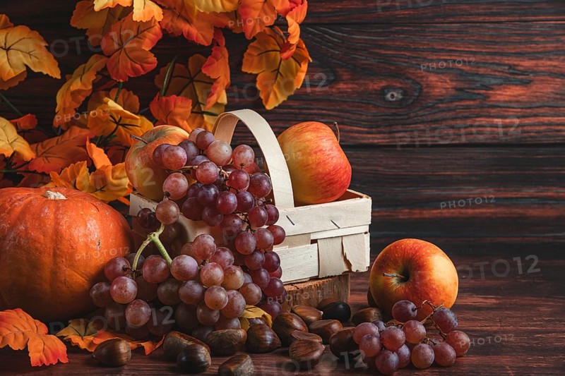 A wooden basket with apples and grapes next to chestnuts, a pumpkin and autumn leaves