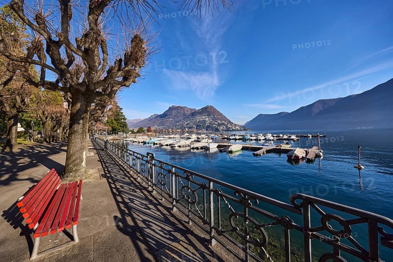 Lake, lakeside promenade, trees, railing, bench, shadow, hilly landscape, marina, motorboats, haze, blue sky, cirrostratus clouds, Lake Lugano, Lugano, district Lugano, canton Ticino, Switzerland, Europe