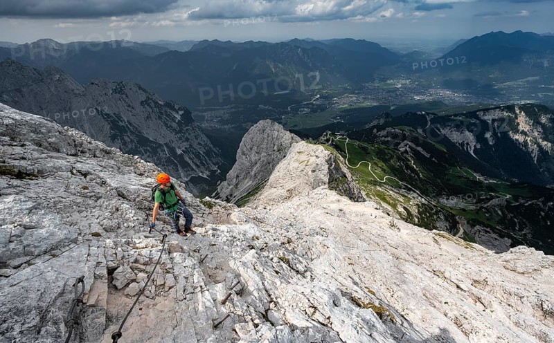 Mountaineer with helmet climbing on the rock in a secured climb, on a narrow rocky ridge, in steep rocky mountain landscape, view of Garmisch Partenkirchen, descent from the Alpsitze via the via ferrata, Alpspitz Ferrata, Wetterstein range, Bavaria, Germany, Europe