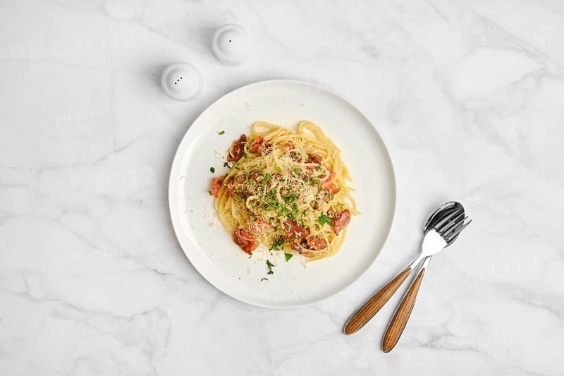 Overhead view of spaghetti carbonara sprinkled with cheese on a marble table. Salt shakers are nearby, enhancing the dining experience with simple elegance