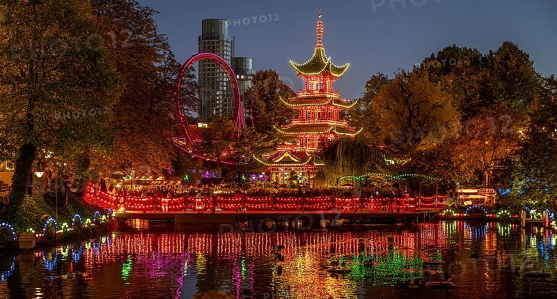 Illuminated pagoda, colourful lighting, roller coaster, trees, cityscape at night with modern building, Tivoli amusement and recreation park, Copenhagen, Denmark, Europe