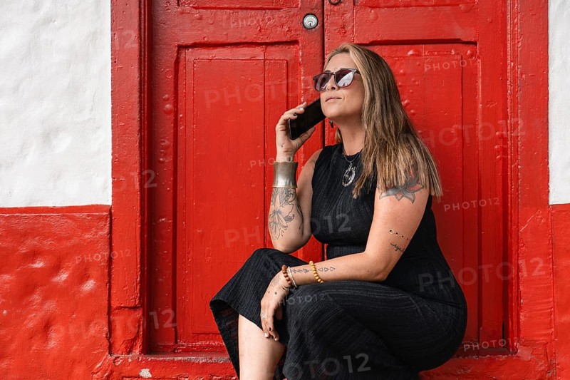 A stylish tattooed woman, dressed in a black dress and sunglasses, holds her smartphone while receiving a call while sitting in front of a red door. A mix of modern lifestyle and urban charm.