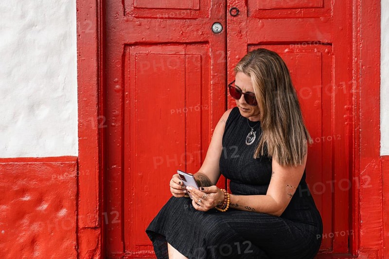 A tattooed woman in sunglasses and black dress is sitting in front of a vibrant red door, concentrating on her smartphone. The urban background adds an elegant and relaxed atmosphere.