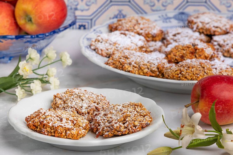 Sugar-dusted oat biscuits on a plate surrounded by apples and flowers in front of patterned tiles