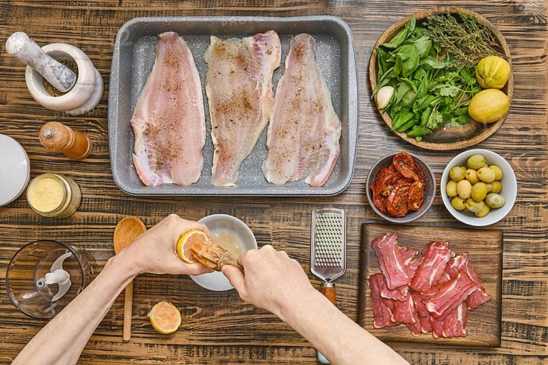 Hands squeeze lemon juice to prepare a marinade for fish fillets arranged in a baking tray