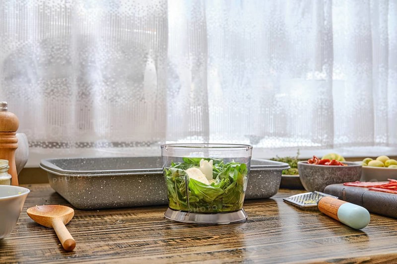 Bright sunlight filters through a window illuminating a kitchen countertop where fresh vegetables are being chopped. Surrounding bowls and tools indicate meal preparation is underway