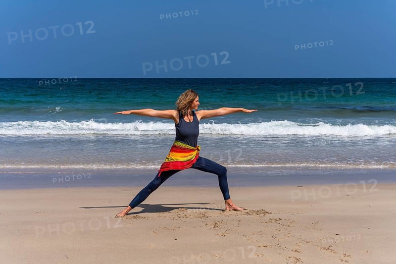 A woman doing yoga on a sandy beach by the sea. Virabhadrasana 2 or Warrior 2. Treen Cove, Cornwall, England, Great Britain
