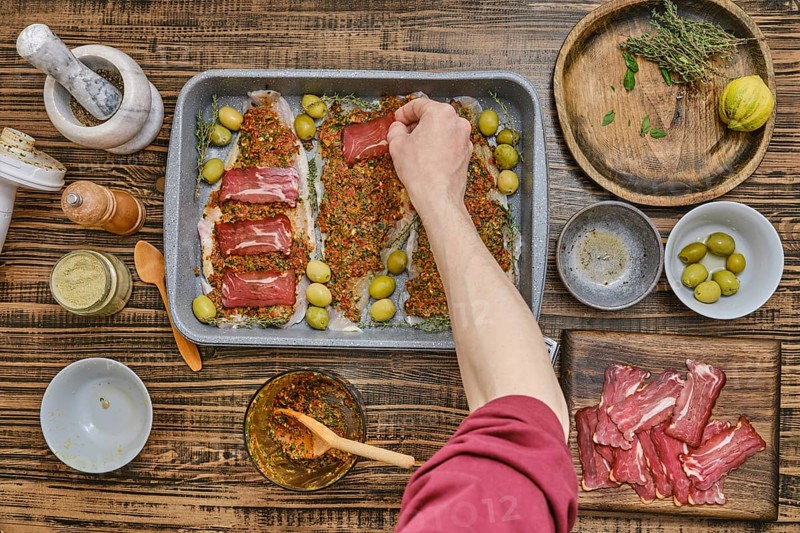 Hand places slices of bacon atop a seasoned fish fillets on a baking tray. Surrounding ingredients include spices and a mortar and pestle on a warm wooden surface