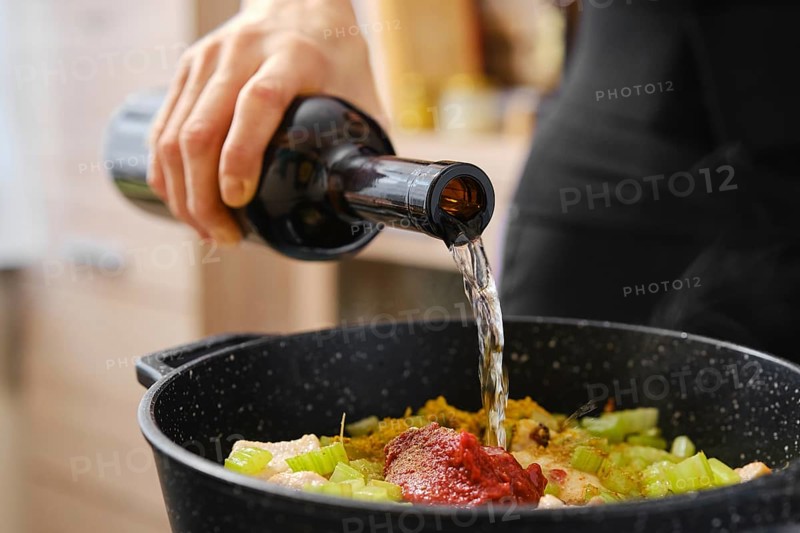 A person pours white wine from a dark bottle into a pot filled with chopped vegetables and seasonings. The setting features a modern kitchen with a focus on the cooking process and fresh ingredients