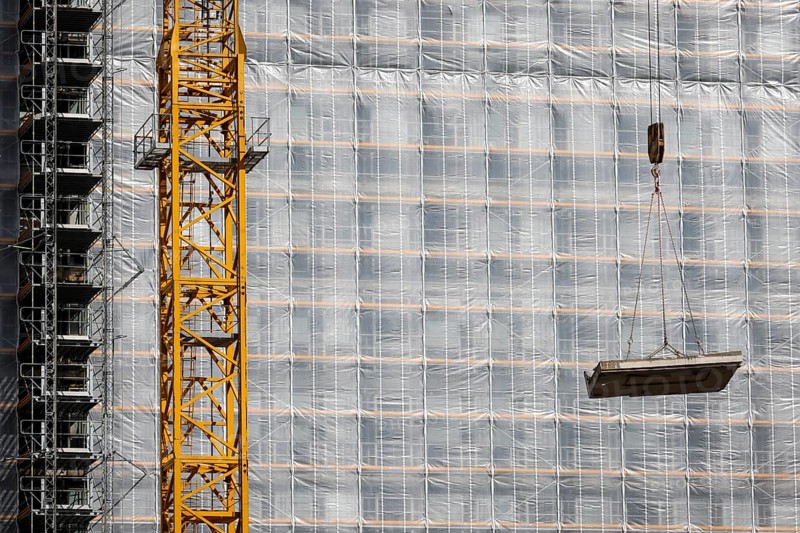 Essen, North Rhine-Westphalia, Germany - Construction crane lifts concrete section in front of scaffolded office building site facade