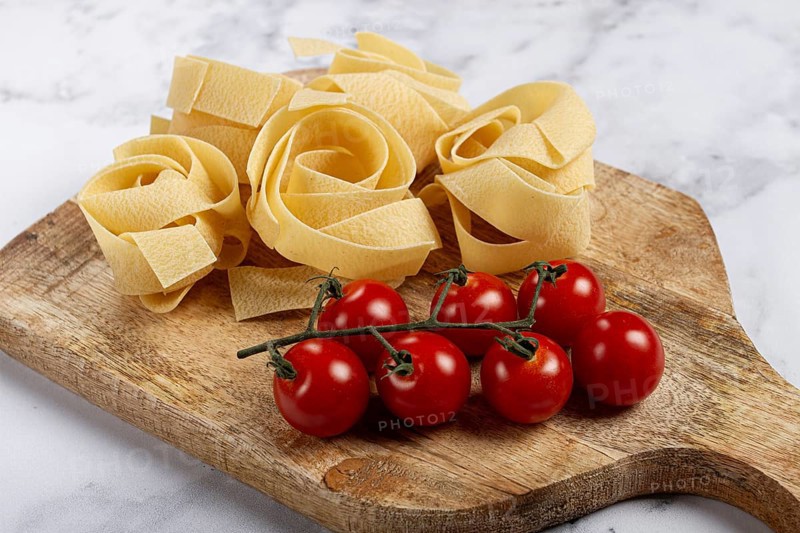 Raw pappardelle pasta, on a wooden chopping board, with cherry tomatoes, top view, no people