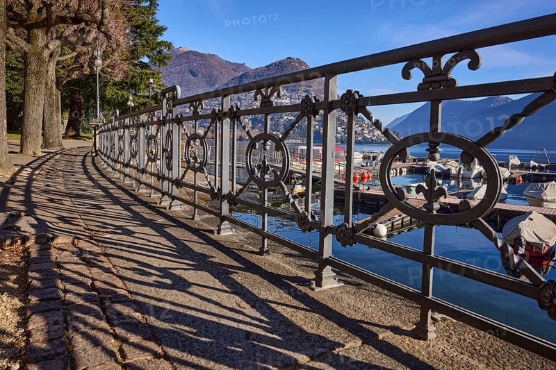 Forged railing with shadow, frog perspective, lakeside promenade, trees, lake, mountains, blue sky, Lake Lugano, Lugano, district of Lugano, canton of Ticino, Switzerland, Europe