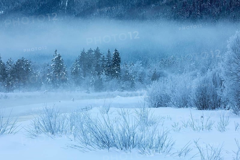 Isar valley in winter at temperatures of minus 18 degrees Celsius. Snow covered landscape with the Isar river flowing through. Some hoar frosted trees and fog. Early morning at sunrise