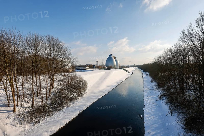 Dortmund, North Rhine-Westphalia, Germany - Sunny winter landscape in the Ruhr area, ice and snow on the renaturalised Emscher, digestion towers at the Dortmund Deusen sewage treatment plant, the treatment plant is now being equipped with a 4th purification stage, which also removes drug residues from the treated wastewater, the ecological conversion of the Emscher system consists of the construction of a central wastewater treatment system in the Ruhr area, the construction of underground sewers and the renaturation of the Emscher and its tributaries
