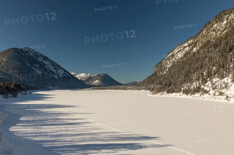 Frozen Sylvenstein reservoir, covered with snow. Isar valley in winter at temperatures of minus 18 degrees Celsius. Snow covered landscape with the Isar river flowing through. Some hoar frosted trees. Early morning at sunrise