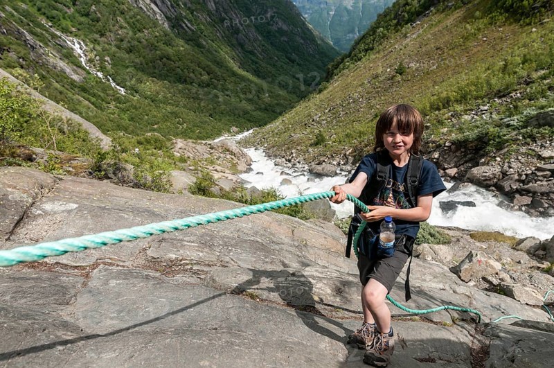 Boy uses rope at steep passage, rock, hiking path to glacier Buarbreen, river in valley stream down to fjord in the background, near Odda, Hardanger, Norway, Europe