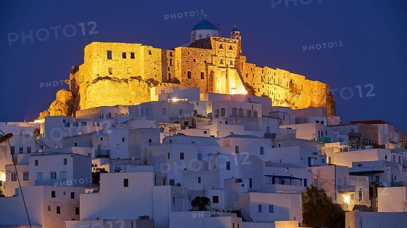 An illuminated historic building towers over plain white structures in the darkness, Querini Castle, Chora, Astypalea, Dodecanese, Greek Islands, Greece, Europe