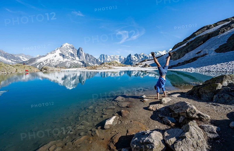 Young man doing a handstand, in front of mountain landscape with mountain lake, tourist at Lac Blanc with water reflection, mountain peaks Aiguille Verte, Grandes Jorasses, Aiguille du Moine and Mont Blanc, Mont Blanc massif, Chamonix-Mont-Blanc, Haute-Savoie, France, Europe
