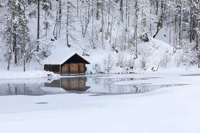 Snow covered boat shelter on Lake Almsee which is partly frozen and snow covered. Snow on the forest with gray sky