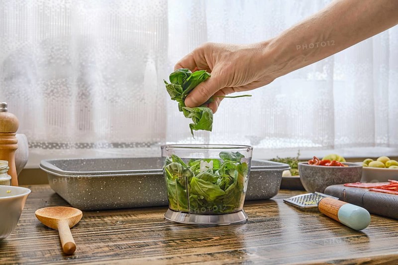 A hand gently places fresh basil leaves into a bowl for salad preparation. The kitchen features wood accents, various ingredients, and bright, natural light creates a warm atmosphere
