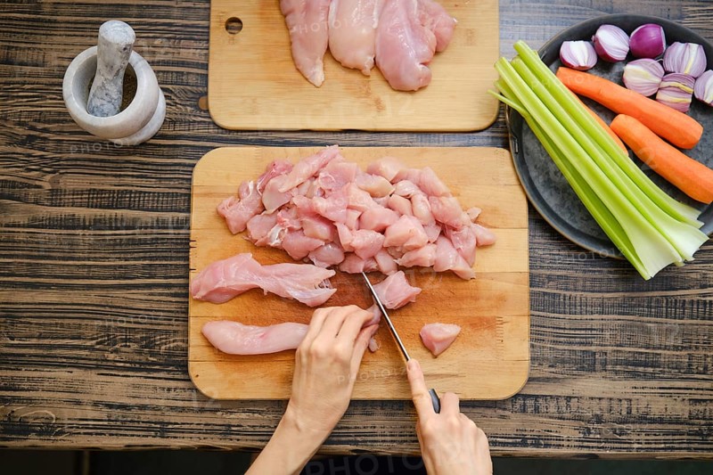 A person skillfully chops raw chicken on a wooden cutting board, surrounded by colorful vegetables like carrots and celery. Ingredients are neatly arranged, enhancing the cooking atmosphere