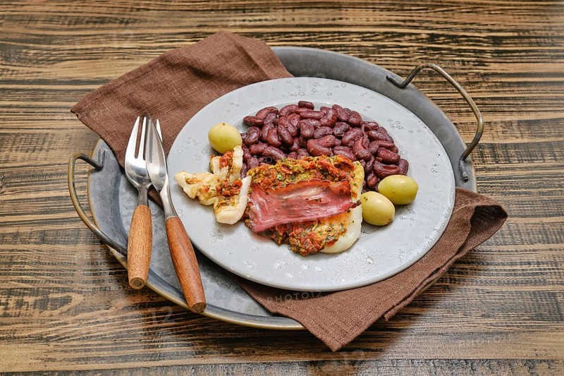 A plate showcases seasoned fish topped with ham, accompanied by red beans and green olives, elegantly arranged on a rustic wooden table with cutlery nearby