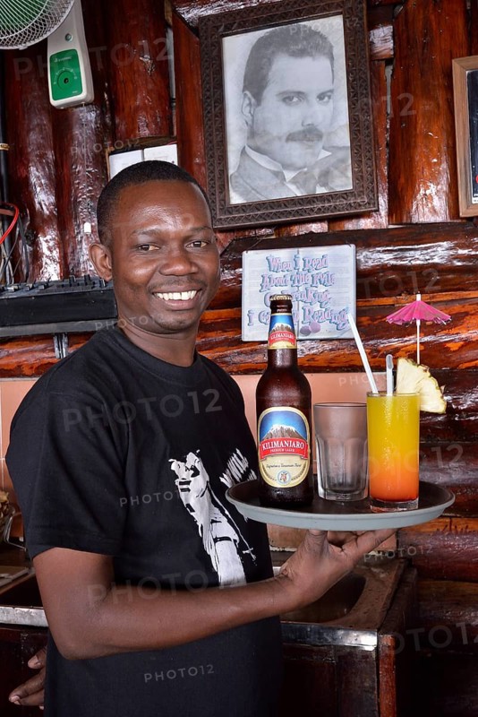 Waiter with Kilimanjaro beer and a cocktail in front of a picture of Freddy Mercury, the late singer of the rock band Queen who was born in Zanzibar, Mercury's Restaurant, Stone Town, Zanzibar City, Zanzibar, Tanzania, Africa