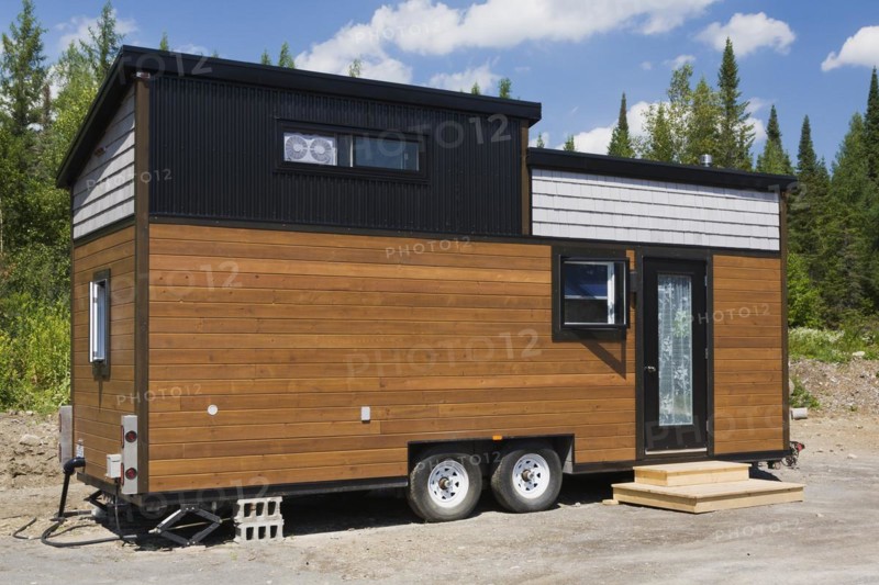 Contemporary brown stained pine and grey oak wood shingles clad cottage style 8 x 24 foot sloped roof mobile Mini House facade with black trimmed windows and entrance door in summer, Quebec, Canada, North America