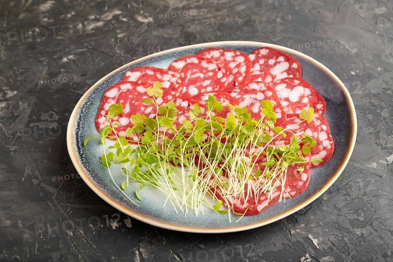 Slices of smoked cervelat salami sausage with spinach microgreen on black concrete background. Side view, close up