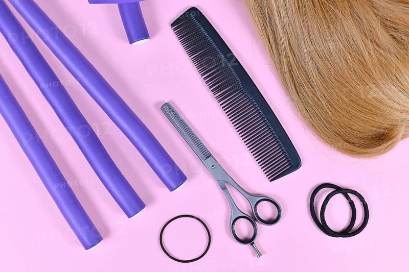 Hair styling concept with dark blond hair, elastic hair ties, comb, thinning shears, and papillote curlers on pink background