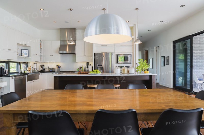 Open-concept dining and kitchen area with antique dining table with black leather and plastic chairs, marble top wooden island with painted black steel and exotic wood bar stools, white cabinets inside a luxurious modern cube style house, Quebec, Canada, North America