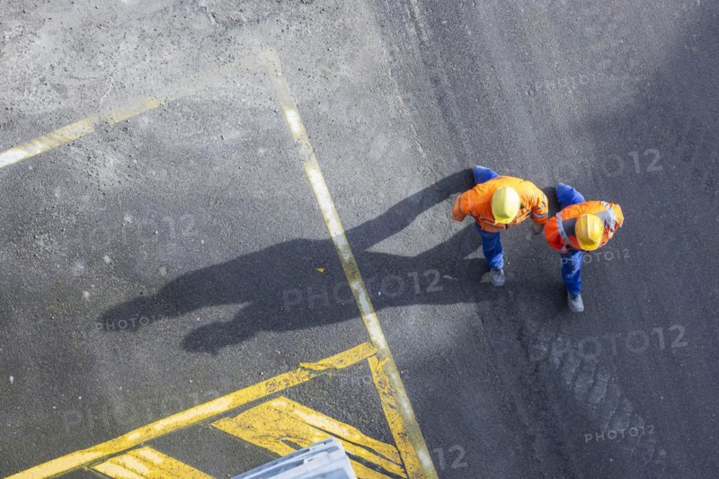 Two construction workers in hard hats walk over tarmac. They cast long shadows. Stuttgart, Baden-Württemberg, Germany, Europe