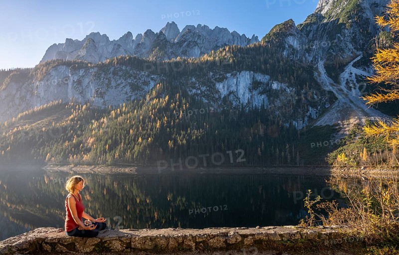 A woman doing yoga by the lake. Lotus position (Padmasana) . Vorderer Gosausee in autumn with a view of the Dachstein mountains. The Gosaukamm on the right. Blue sky, good weather. Reflection. Morning atmosphere. Vorderer Gosausee, Gosau, Gosau valley, Salzkammergut, Upper Austria, Austria, Europe