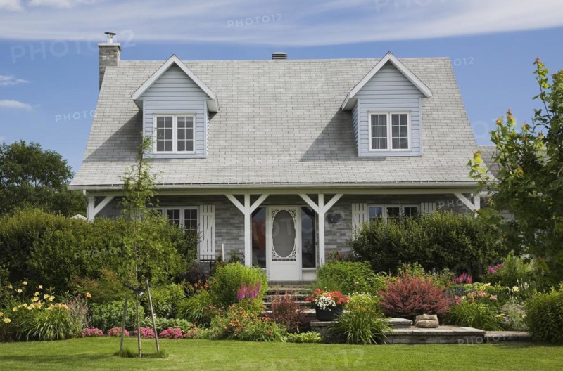 Facade of old grey nuanced brick with white trim cottage style Canadiana home with dormer windows and landscaped front yard with mixed perennial shrubs and flowers in borders in summer, Quebec, Canada, North America