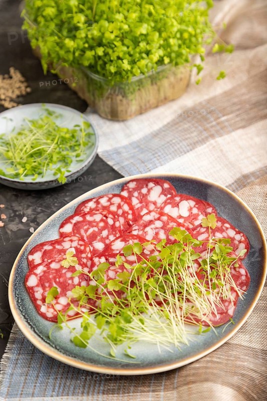 Slices of smoked cervelat salami sausage with spinach microgreen, salt and pepper on black concrete background and beige textile. Side view, close up, selective focus