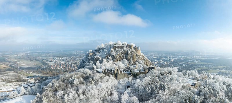 Aerial panorama of the snow-covered Hegau volcano Hohentwiel with Germany's largest castle ruins, surrounded by frosty trees in a winter landscape, with the town of Singen, district of Constance, Baden-Württemberg, Germany in the background