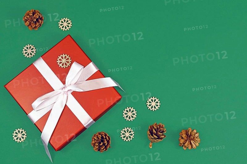 Red Christmas gift box with ribbon surrounded by seasonal pine cones and wooden snowflake ornaments in corner of green background with empty copy space