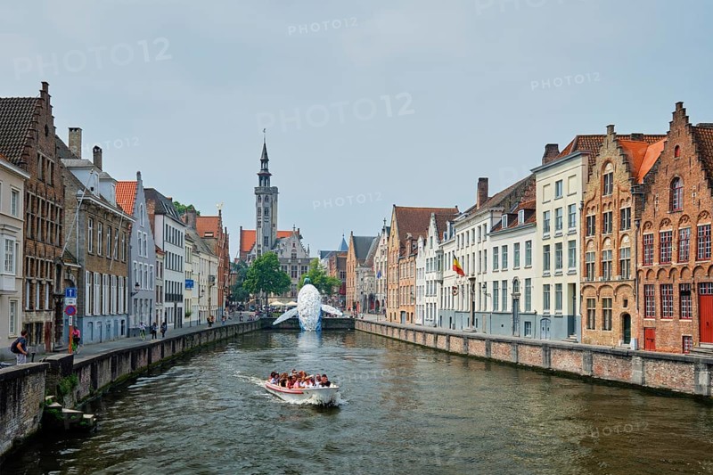 BRUGES, BELGIUM, MAY 29, 2018: Tourist boat in canal between old houses with The Bruges Whale plastic installation and The Burghers' Lodge Poortersloge tower. Brugge Bruges, Belgium, Europe