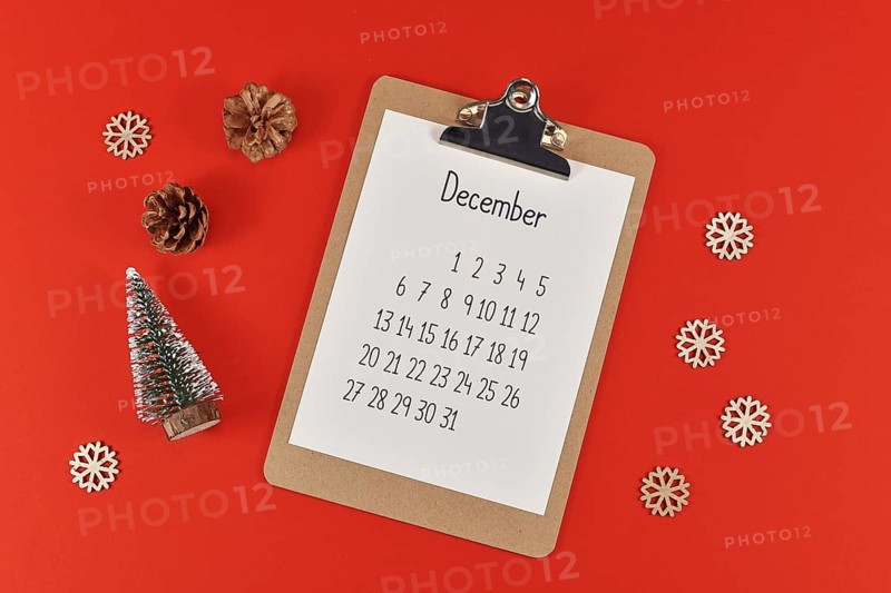 Christmas flat lay with clipboard with calendar sheet for month December surrounded by small Christmas trees, pine cones, snowballs and snowflake ornaments on red background