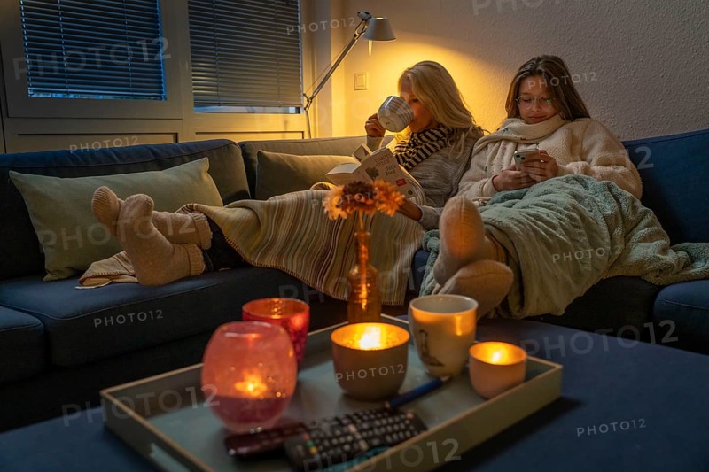Symbolic image energy saving, cold winter, energy crisis, cold flat, mother and daughter lying on the sofa, warmly dressed, candlelight, heating on to a minimum