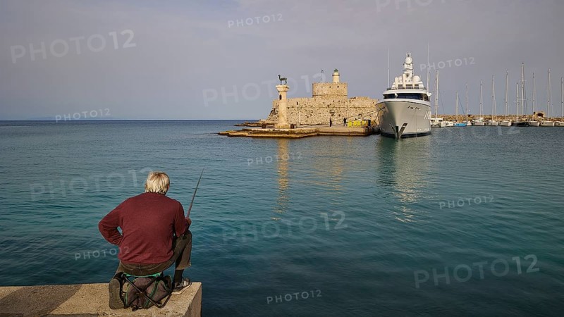 A man fishing on a shore with a view of an old fortress and a luxury yacht at sunset, Fort of Saint Nikolaos, harbour area, Rhodes Town, Rhodes, Dodecanese, Greek Islands, Greece, Europe