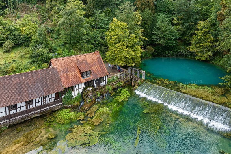 Blautopf Blaubeuren with industrial monument Hammerschmiede, source of the little river Blau in a landscape with forest. Karst spring, geotope and geopoint of the UNESCO Swabian Alb Geopark, tourist attraction. The popular excursion destination is now being thoroughly renovated and will therefore be closed to visitors until the end of 2028. Drone photo. Blaubeuren, Baden-Württemberg, Germany, Europe