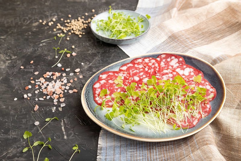 Slices of smoked cervelat salami sausage with spinach microgreen, salt and pepper on black concrete background and beige textile. Side view, close up