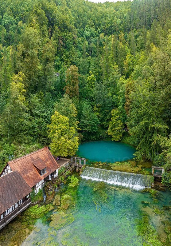 Blautopf Blaubeuren with industrial monument Hammerschmiede, source of the little river Blau in a landscape with forest. Karst spring, geotope and geopoint of the UNESCO Swabian Alb Geopark, tourist attraction. The popular excursion destination is now being thoroughly renovated and will therefore be closed to visitors until the end of 2028. Drone photo. Blaubeuren, Baden-Württemberg, Germany, Europe