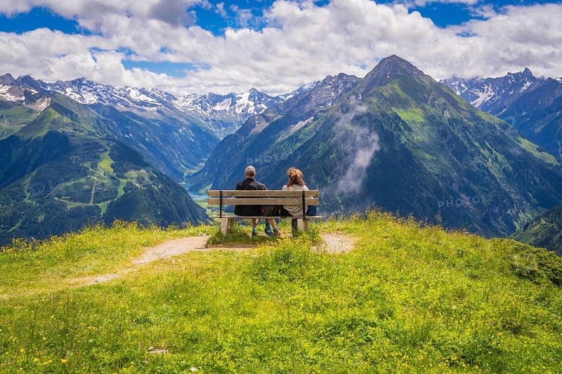 Two people sit on a bench and look out over a valley with majestic mountains in the background, Penken, Zillertal. Austria
