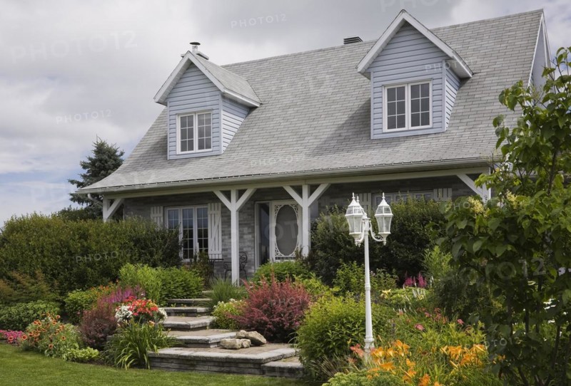 Facade of old grey nuanced brick with white trim cottage style Canadiana home with dormer windows and landscaped front yard with mixed perennial shrubs and flowers in borders in summer, Quebec, Canada, North America