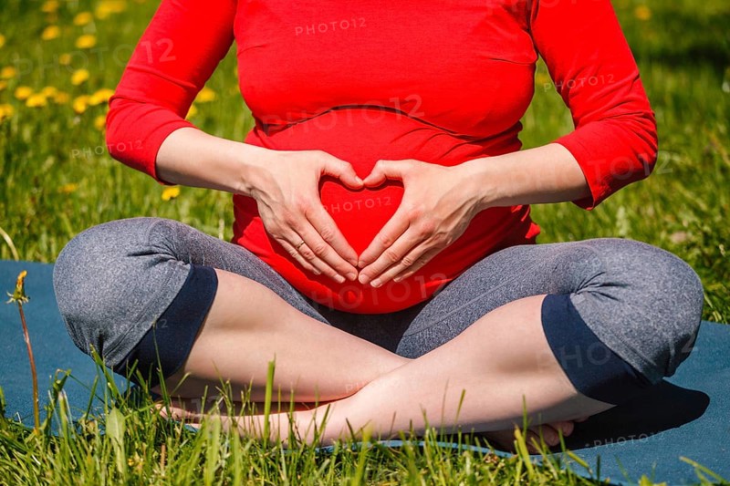 Pregnancy yoga exercise, pregnant woman doing asana Sukhasana easy yoga pose showing heart symbol with hands outdoors on grass lawn with dandelions in summer